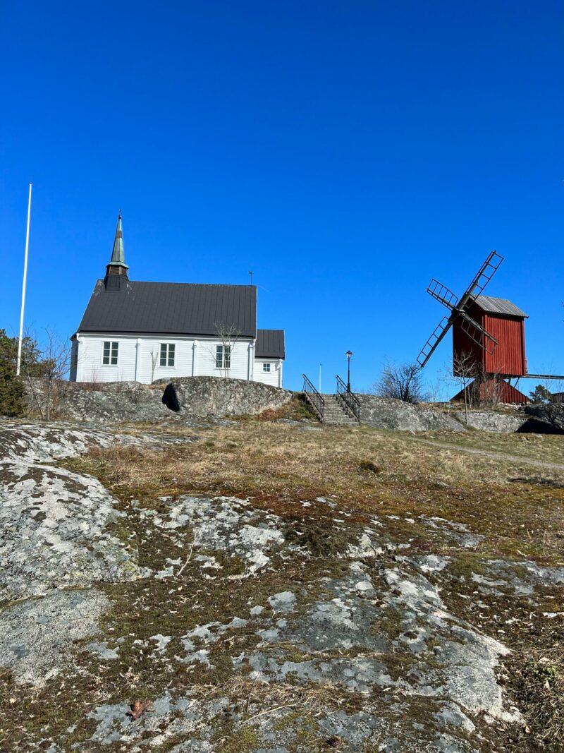 White wooden church and historic red windmill on rocky hilltop under clear blue sky on the Stockholm Archipelago Trail